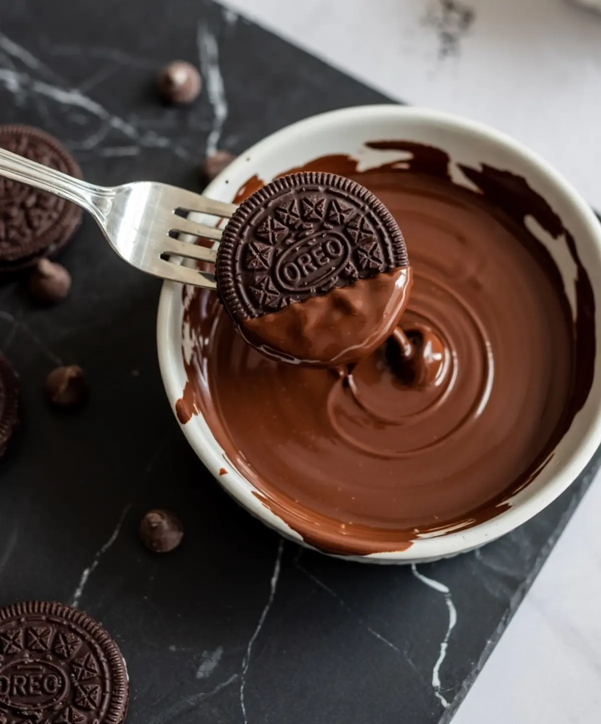 A chocolate sandwich cookie held by a fork being dipped halfway into a bowl of melted milk chocolate, surrounded by extra cookies and chocolate chips on a black marble board.