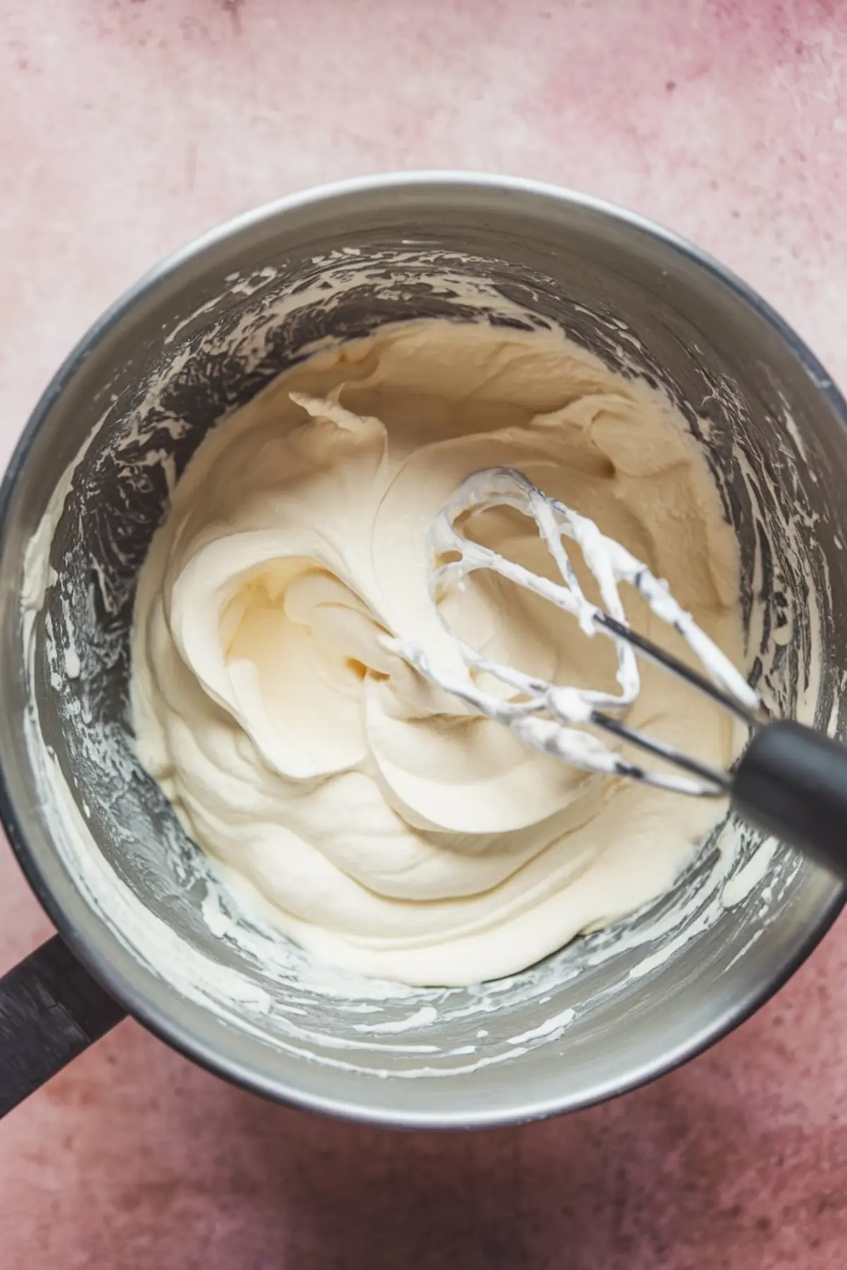 Thick whipped cream being mixed in a metal bowl with an electric hand mixer, creating soft, fluffy peaks.
