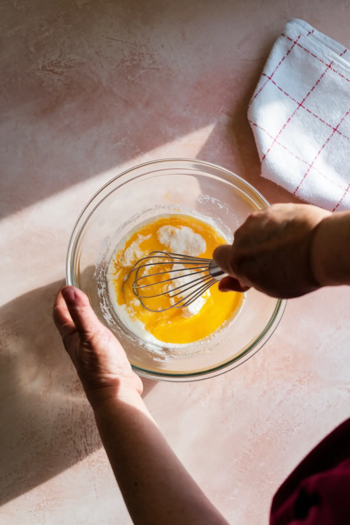 A person whisking egg yolks, sugar, and flour in a clear glass mixing bowl on a pink countertop with natural sunlight and a white kitchen towel nearby.