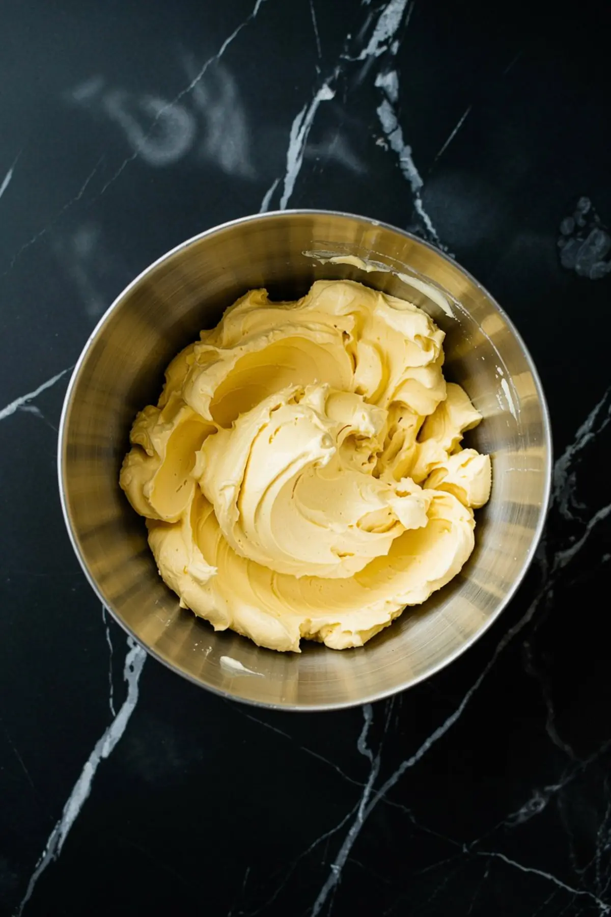 Creamy whipped butter in a stainless steel mixing bowl placed on a black marble surface, ready for chocolate frosting preparation.
