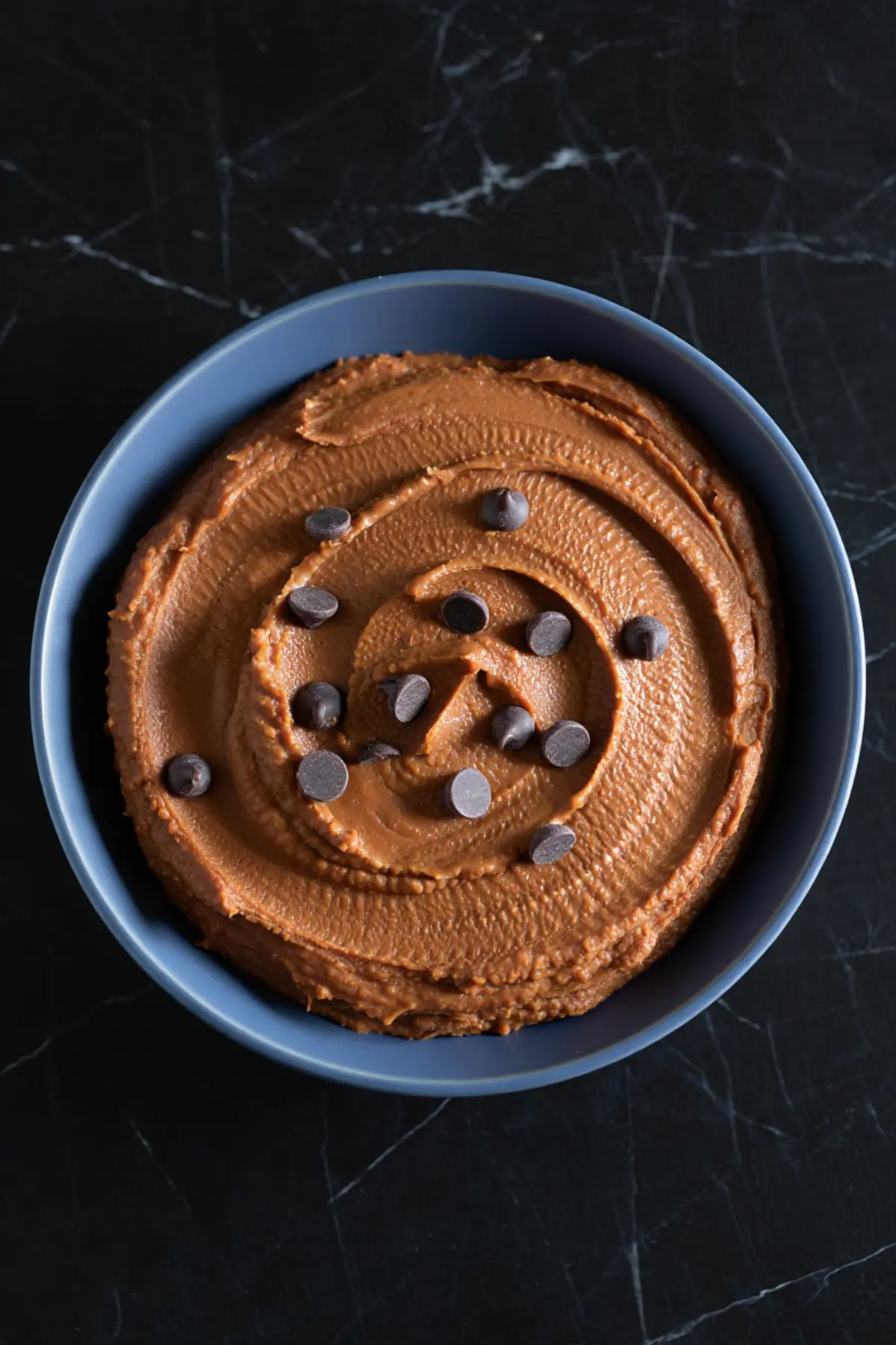 Smooth chocolate hummus topped with dark chocolate chips served in a matte blue bowl on a black marble surface.

