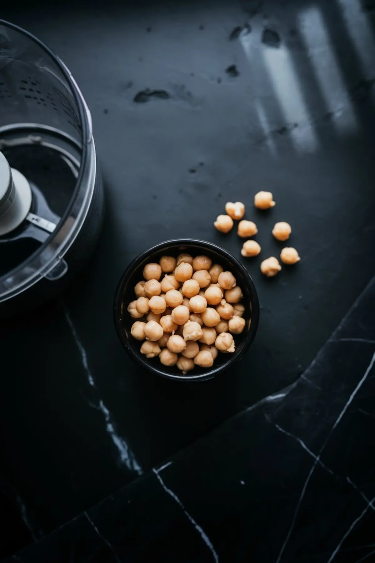 Bowl of canned chickpeas with several scattered on the side, placed next to a food processor on a black marble surface.