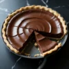 Chocolate pie with a glossy, rippled ganache surface and a golden pastry crust, partially sliced to reveal a smooth and creamy interior on a black marble surface.