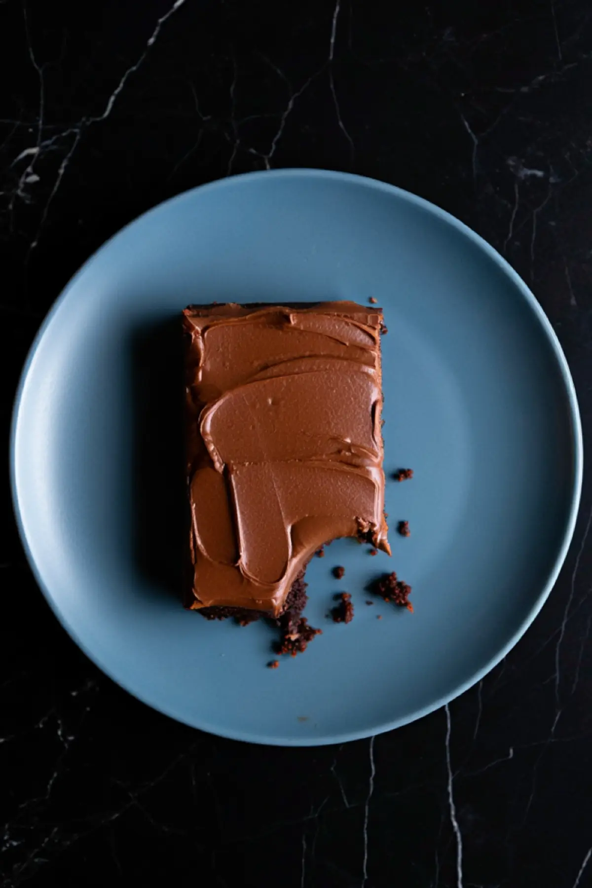 Overhead view of a bitten chocolate sheet cake slice on a blue plate, with scattered cake crumbs highlighting the soft texture.
