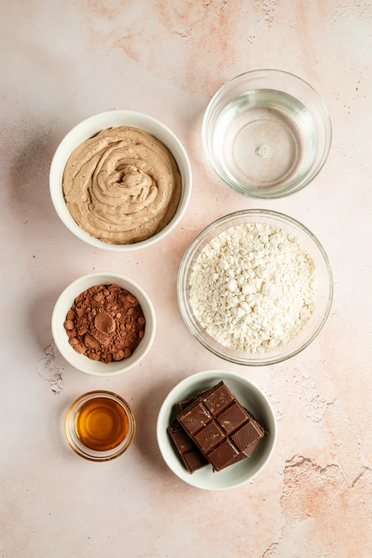 Overhead view of chocolate sourdough bread ingredients arranged on a light pink surface, including all-purpose flour, cocoa powder, sourdough starter, water, chopped dark chocolate, vanilla extract, and a creamy base.