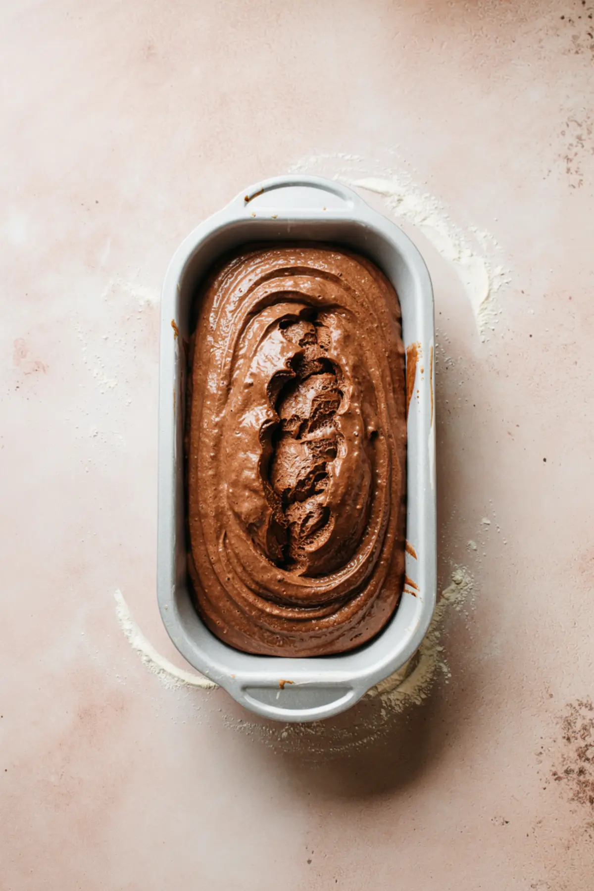 Loaf pan filled with unbaked chocolate sourdough batter featuring a smooth, swirled top on a floured pink background.