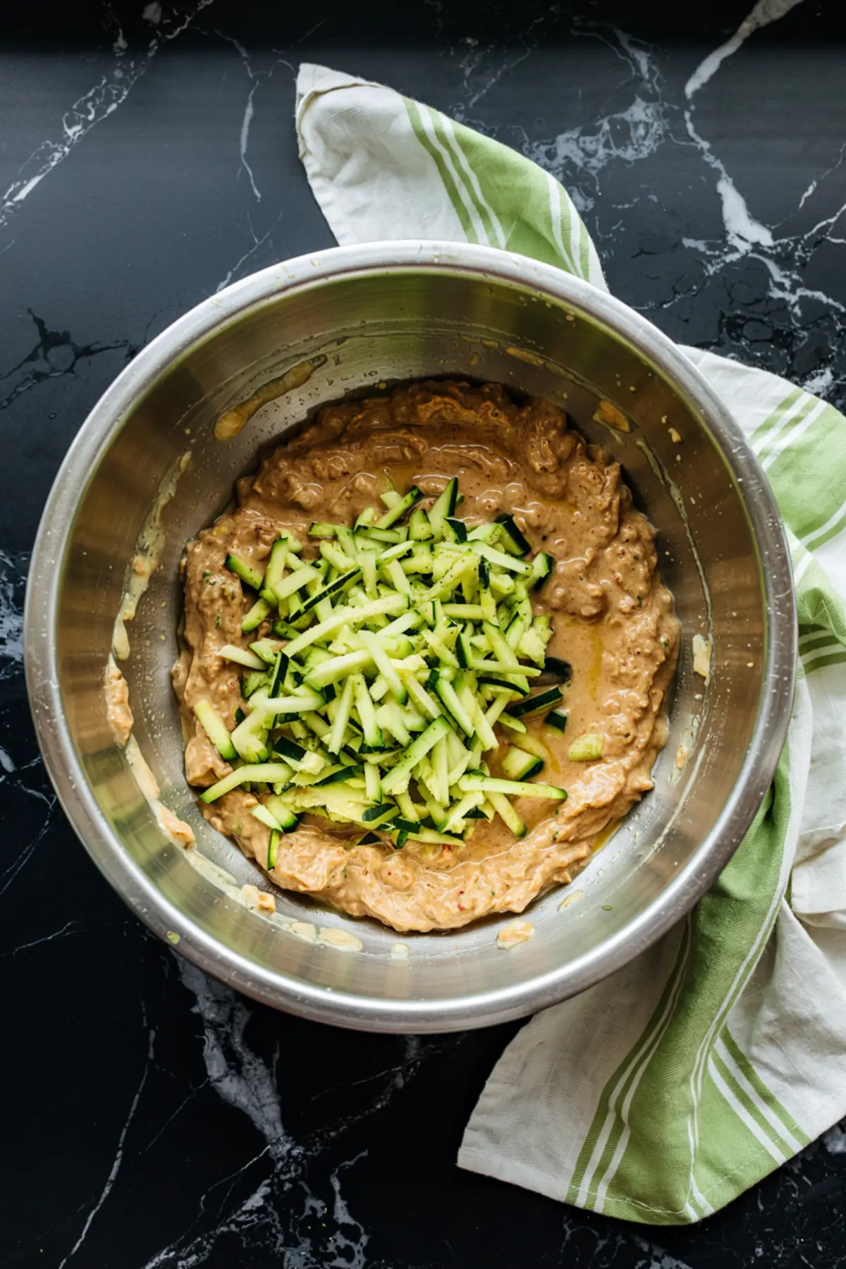 Bowl of zucchini bread batter with freshly shredded zucchini in the center, placed on a black marble surface with a green-striped dish towel.