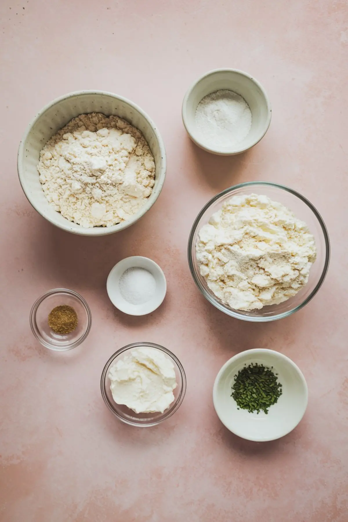 Overhead view of separate baking ingredients in small bowls, including flour, baking powder, salt, cottage cheese, dried herbs, and a seasoning mix, arranged on a pink background.