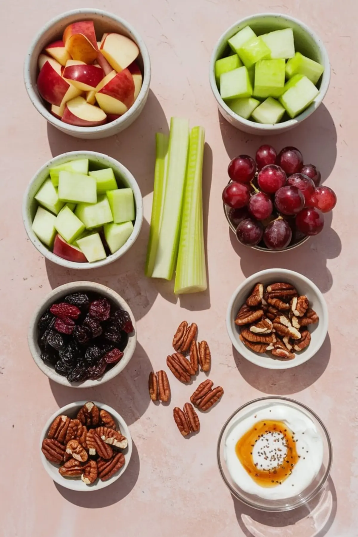 Flat lay of apple salad ingredients including red apple slices, green apple chunks, celery sticks, red grapes, dried cranberries, whole and chopped pecans, and a bowl of yogurt with honey and poppy seeds, arranged neatly on a pink surface.