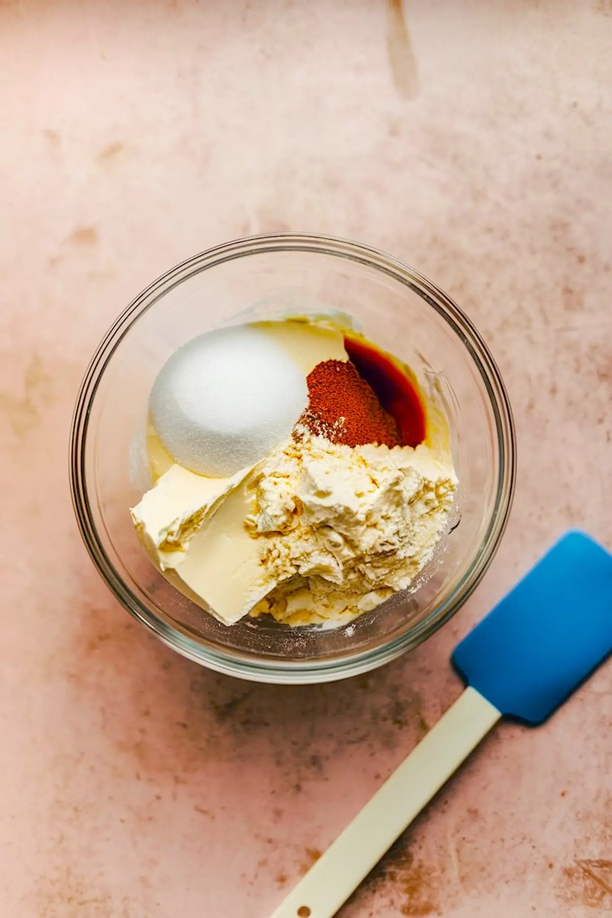 Top-down view of a glass bowl filled with ingredients for banana muffin cream filling, featuring cream cheese, granulated sugar, vanilla extract, and dry mix, with a blue spatula beside the bowl.