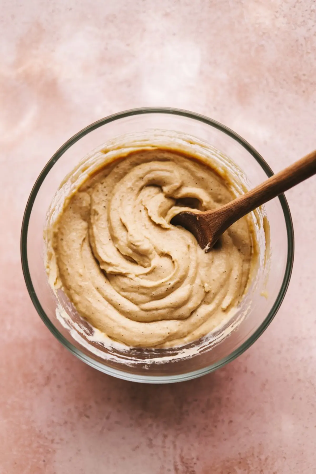 Overhead view of thick banana muffin batter swirled in a glass mixing bowl, with a wooden spoon resting inside, placed on a soft pink countertop.