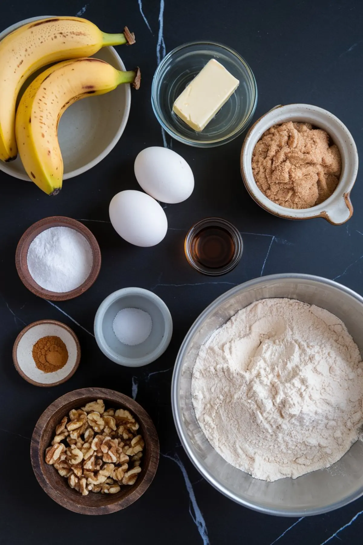 Flat lay of banana bread ingredients arranged on a dark marble surface, including ripe bananas, brown sugar, butter, eggs, flour, baking soda, vanilla extract, cinnamon, salt, and chopped walnuts in ceramic and glass bowls.
