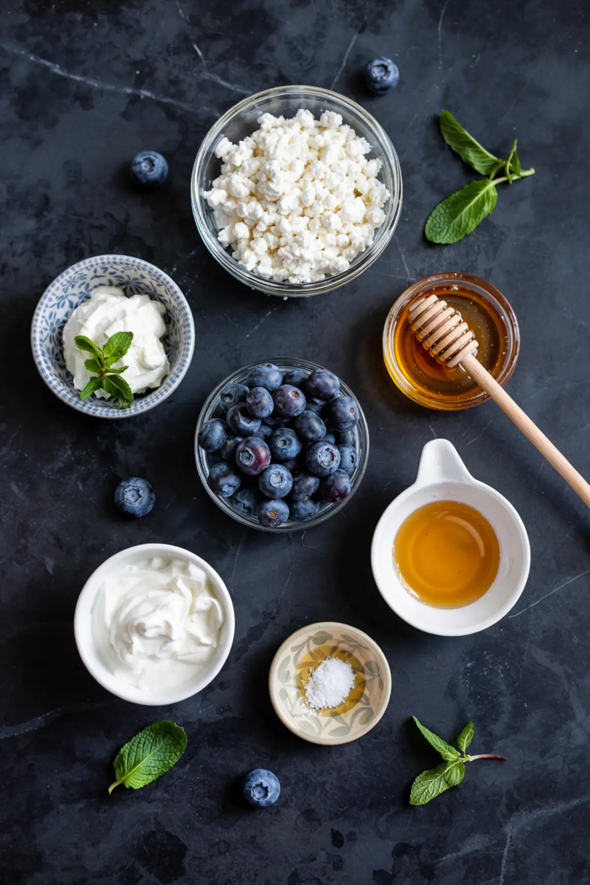Flat lay of ingredients for blueberry cottage cheese dip arranged on a black surface. Includes bowls of cottage cheese, blueberries, honey, Greek yogurt, cream, salt, and garnishes of fresh mint, showcasing the recipe components.