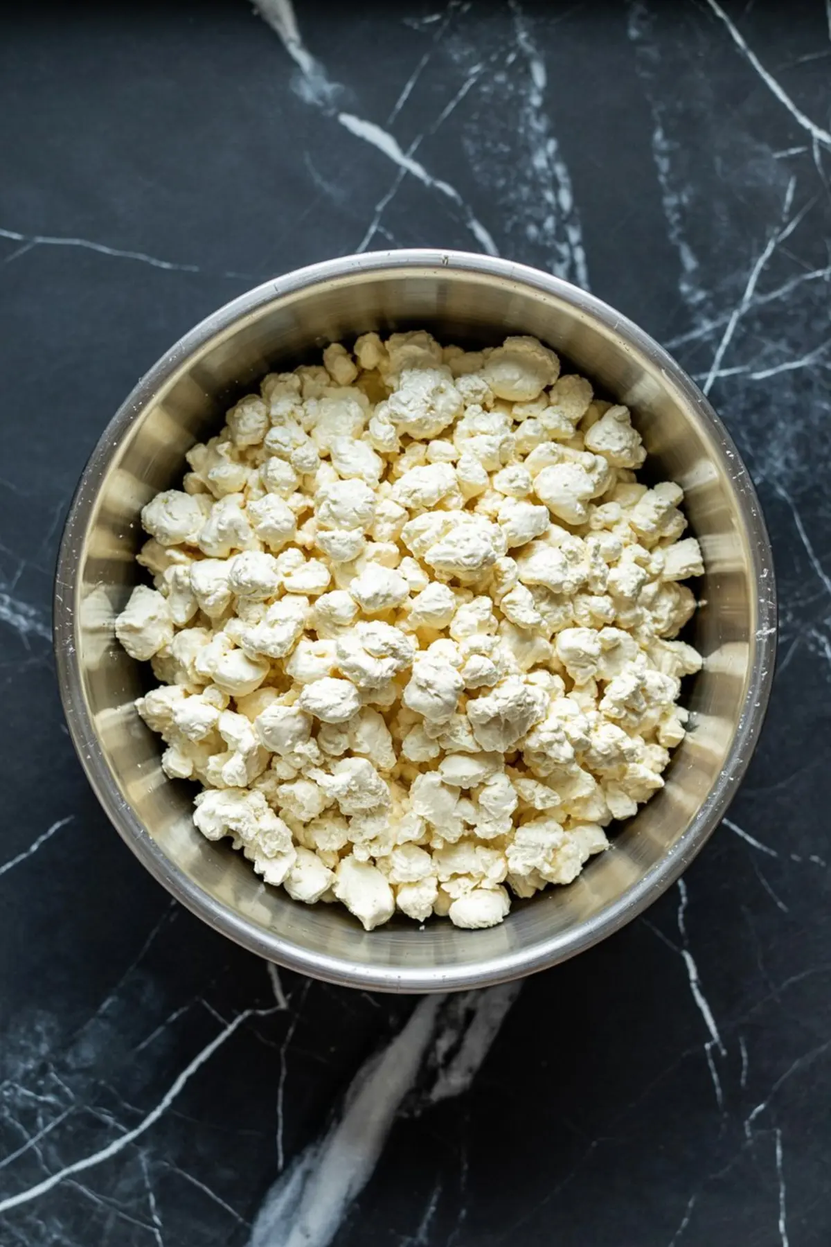Overhead view of a metal mixing bowl filled with chunky cottage cheese, set on a dark marble surface. The textured curds are clearly visible, emphasizing the dairy base for the dip.