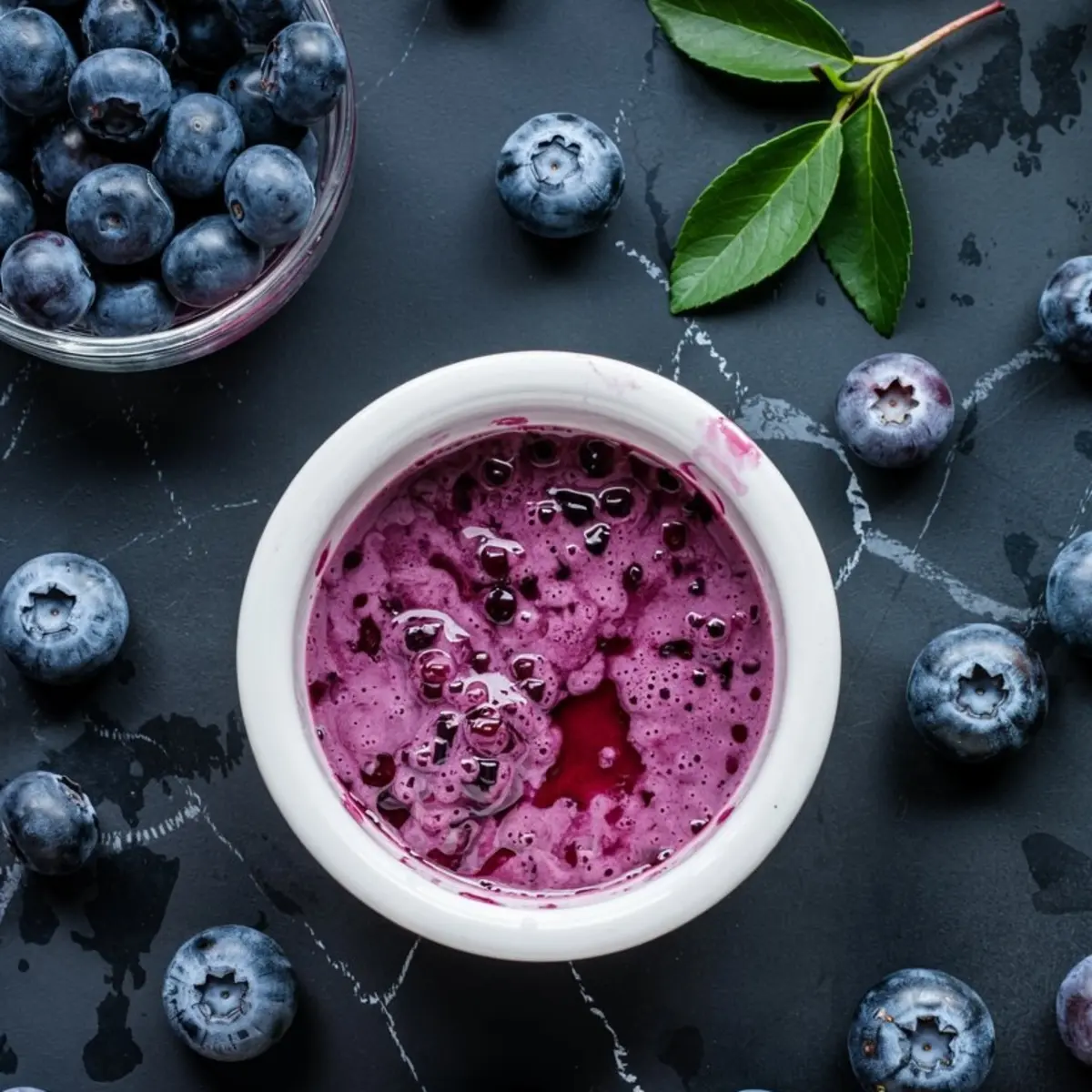 Small white bowl filled with vibrant blueberry puree, surrounded by whole blueberries and a sprig of greenery. The thick, deep purple mixture is highlighted by a glossy surface and visible blueberry skins.