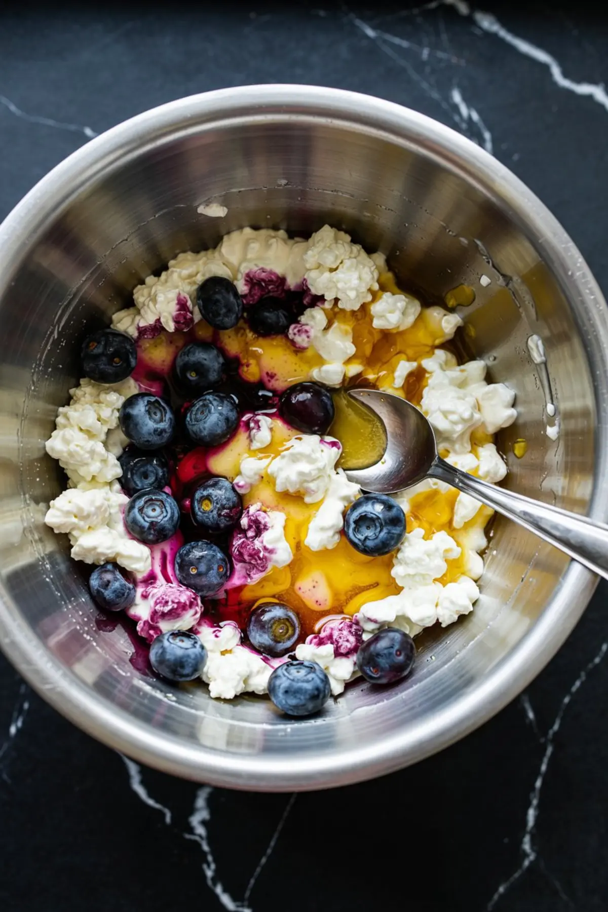 Mixing bowl containing the ingredients for blueberry cottage cheese dip: whole blueberries, golden honey, and creamy cottage cheese. A spoon rests in the bowl, and colorful juice from the berries begins to blend with the other ingredients.