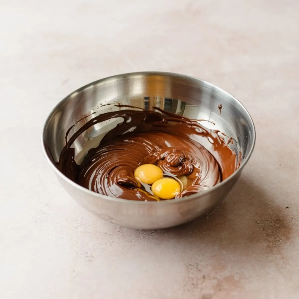 Stainless steel mixing bowl filled with melted chocolate and three raw eggs on a light countertop, showing the early step of making homemade brownie batter.