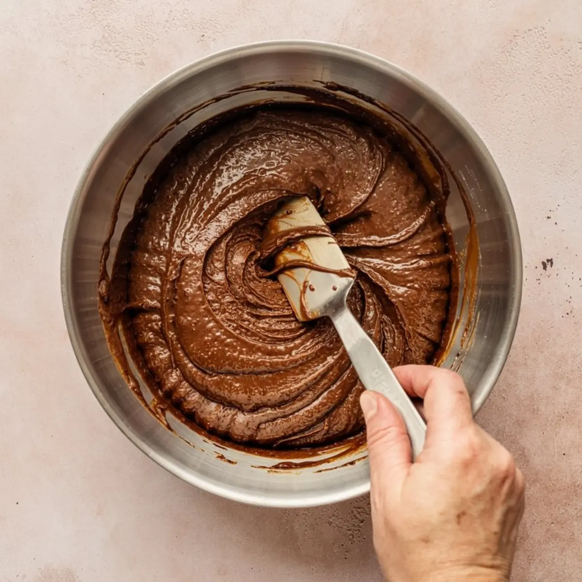 Thick, rich chocolate brownie batter being stirred in a metal bowl with a spatula held by Lena's hand, showing smooth texture and deep color.