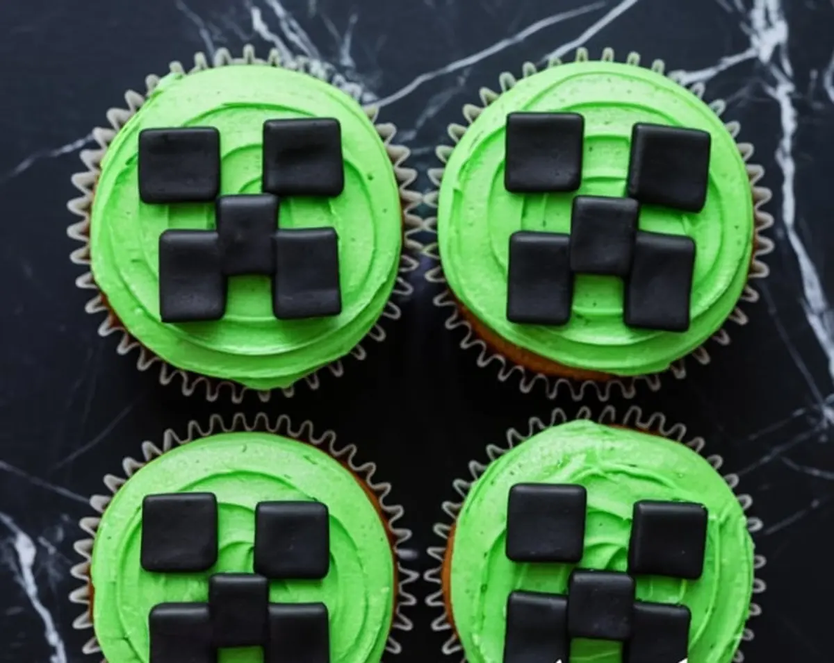 Close-up shot of four chocolate cupcakes topped with green frosting and square black fondant forming Minecraft Creeper faces, displayed on a black marble surface.