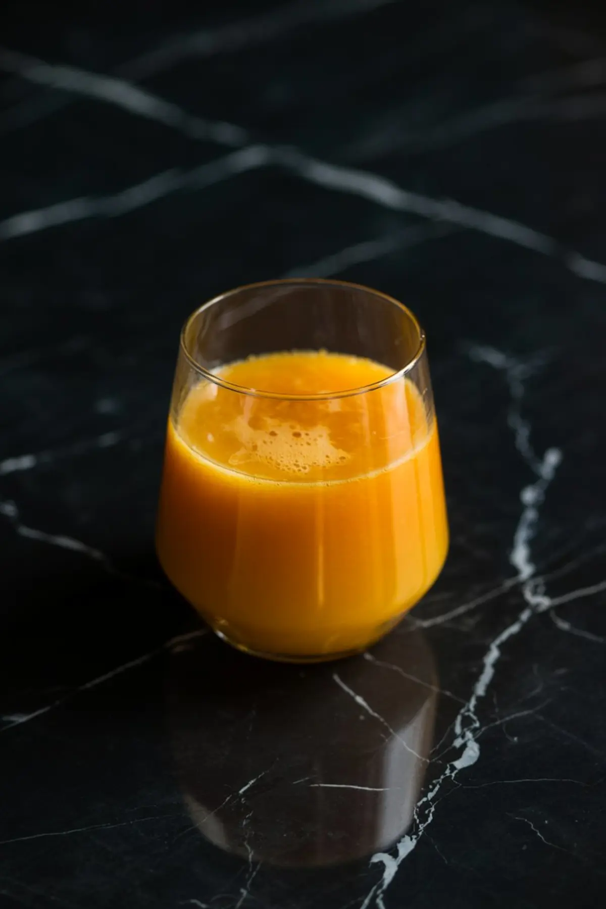 A clear glass filled with vibrant orange juice resting on a sleek black marble table with white streaks, showcasing a bright and fresh citrus drink.
