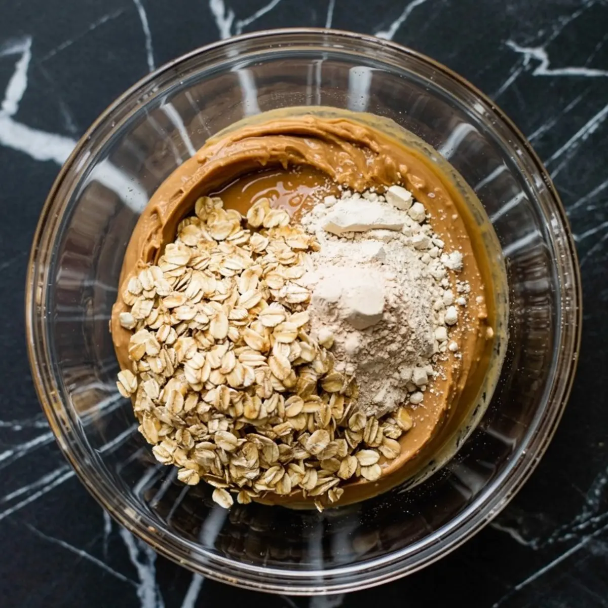 Close-up of a glass bowl filled with oats, peanut butter, and protein powder ready to be mixed for homemade protein balls, placed on a black marble countertop.