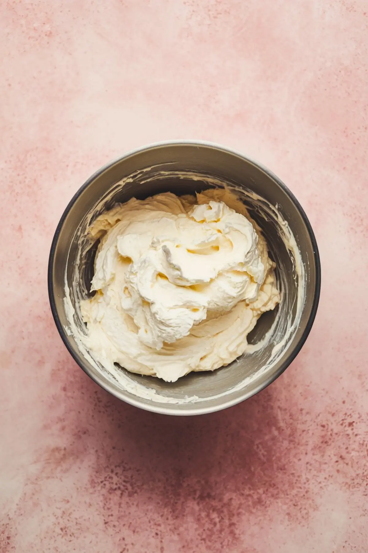Stainless steel mixing bowl filled with freshly whipped cream cheese mixture, resting on a soft pink background. The smooth texture and creamy consistency are ideal for dessert dip preparation.
