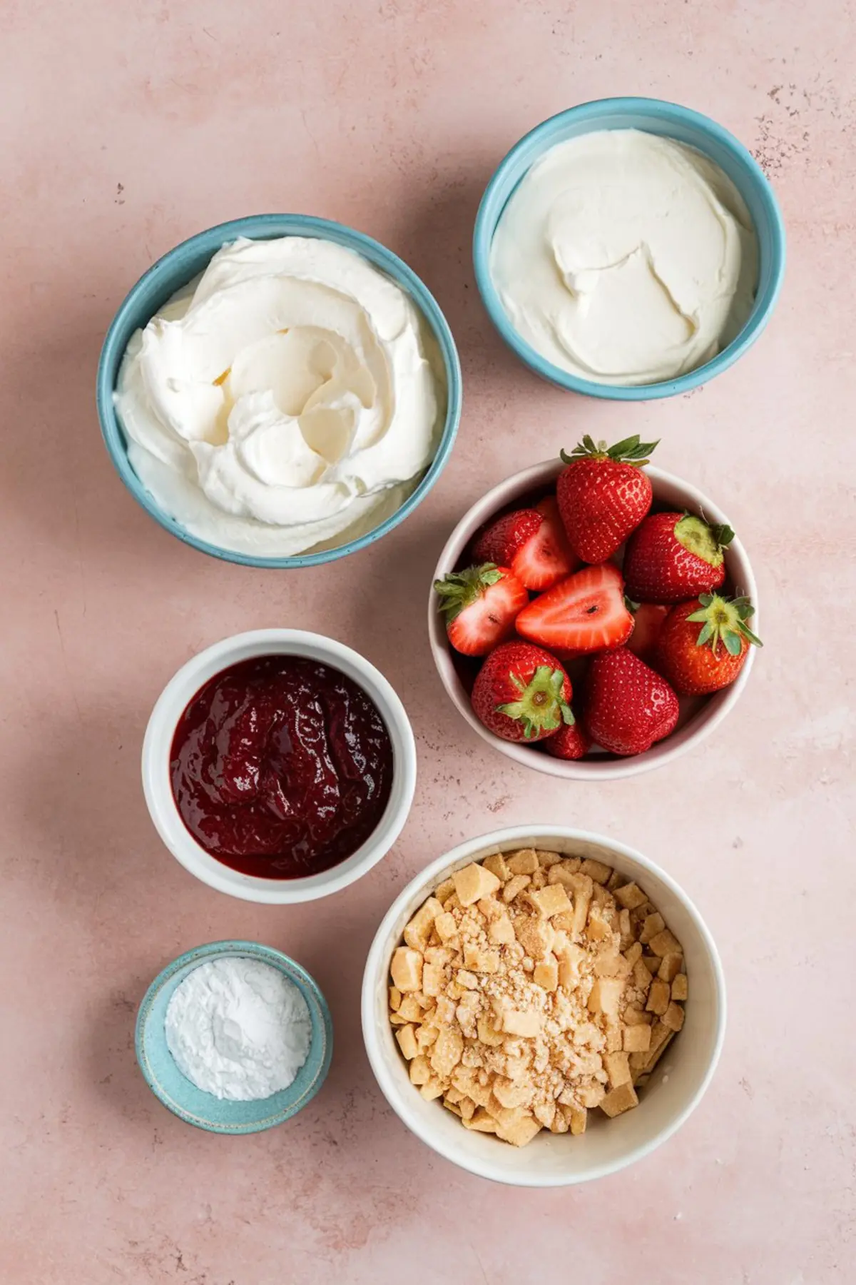 Flat lay of strawberry shortcake dip ingredients on a pink surface, featuring bowls of whipped topping, cream cheese, fresh strawberries, strawberry jam, crushed shortbread cookies, and powdered sugar.
