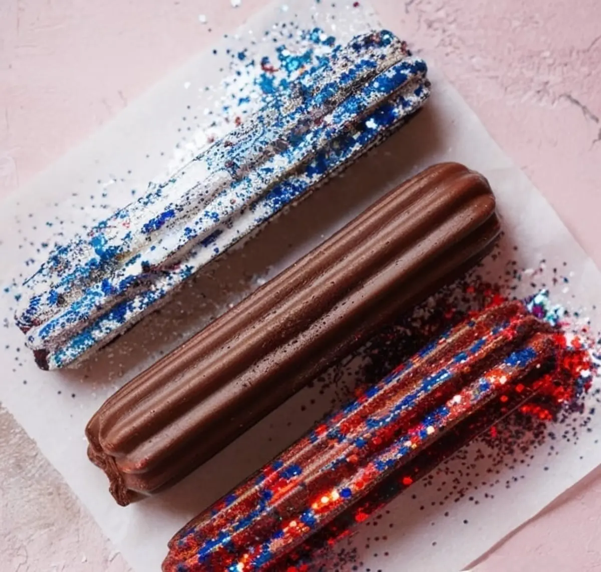 Three chocolate churros on a sheet of parchment paper, two decorated with patriotic blue, red, and silver glitter and one plain, scattered with extra sprinkles on a pink surface.