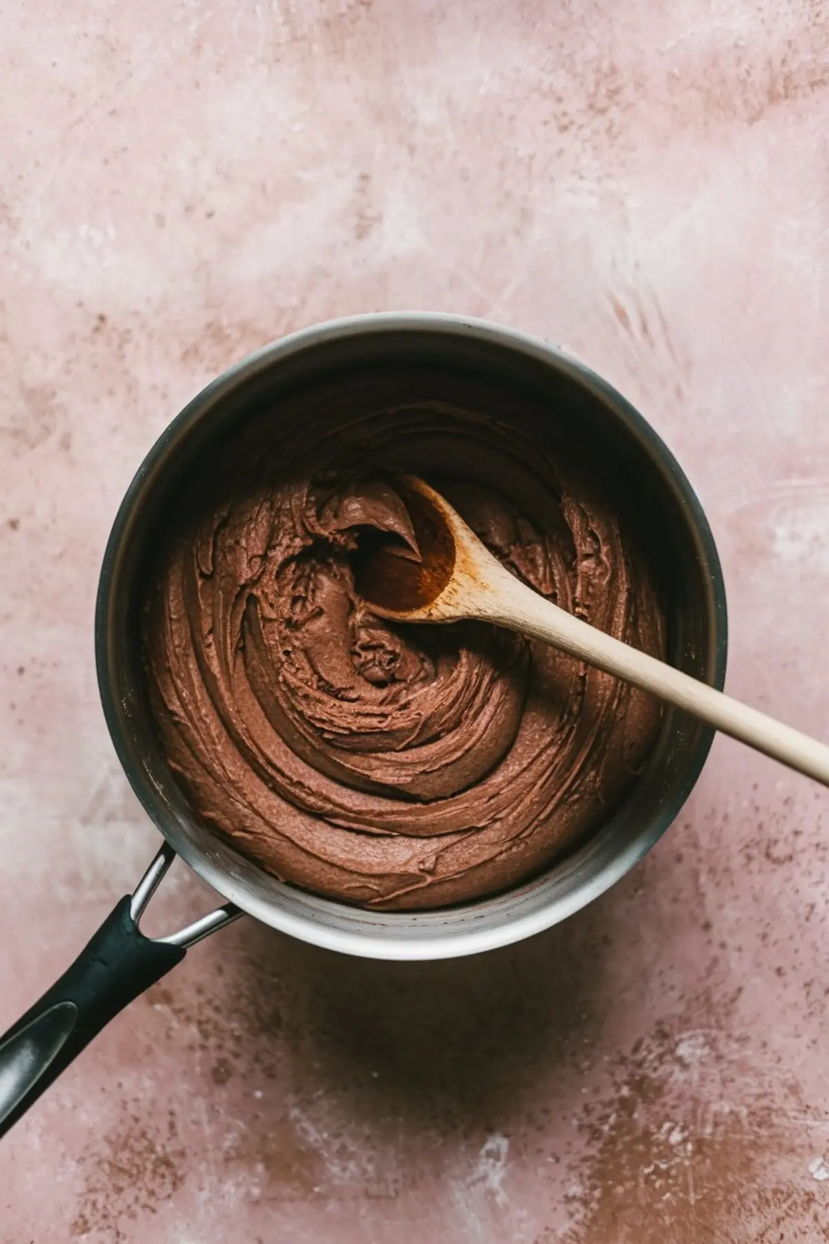 Chocolate churro dough mixed to a smooth, thick consistency inside a saucepan with a wooden spoon, ready for piping and frying, resting on a pink counter.
