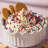 Whipped funfetti dip swirled high with red, white, and blue sprinkles in a white scalloped bowl, garnished with round biscuits and surrounded by graham crackers on a pastel pink background.