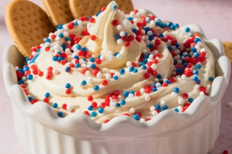 Whipped funfetti dip swirled high with red, white, and blue sprinkles in a white scalloped bowl, garnished with round biscuits and surrounded by graham crackers on a pastel pink background.