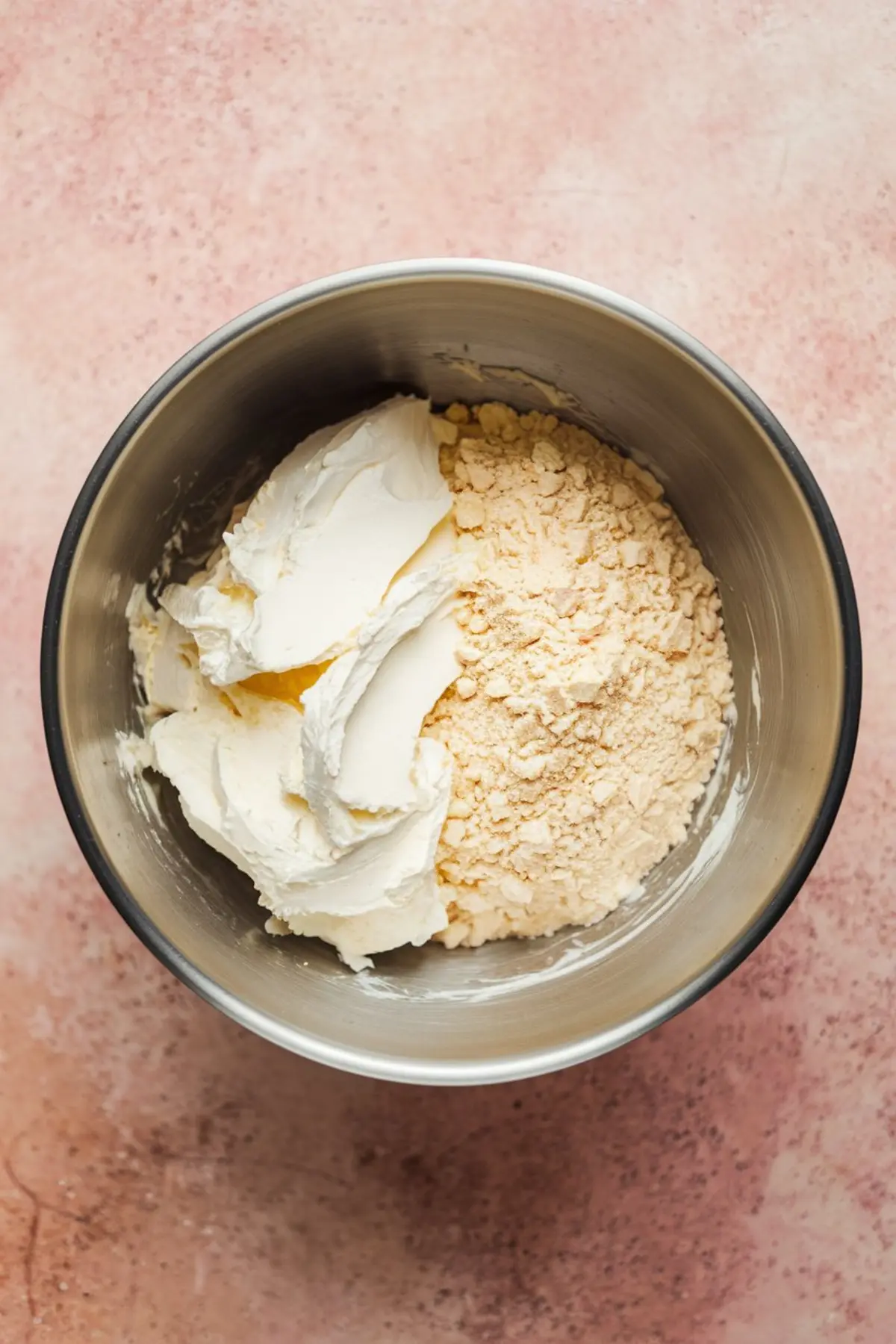 Stainless steel mixing bowl filled with whipped cream cheese and dry cake mix on a pink background, showing the base ingredients for funfetti dip.