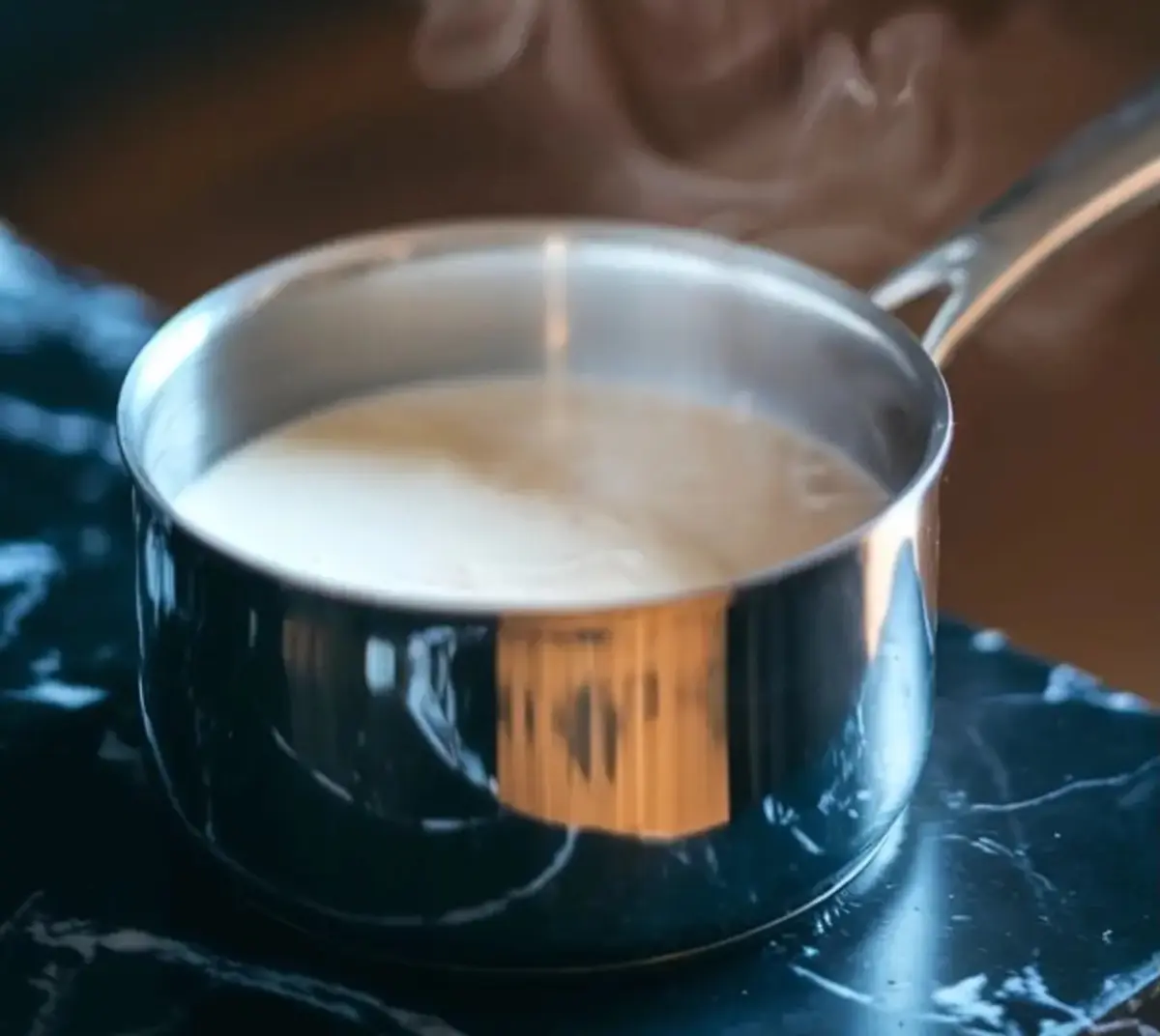 A stainless steel saucepan filled with hot milk, emitting gentle steam, placed on a dark marble counter. Captures the initial step in making homemade hot chocolate, ideal for visual recipe guides and cooking tutorials.