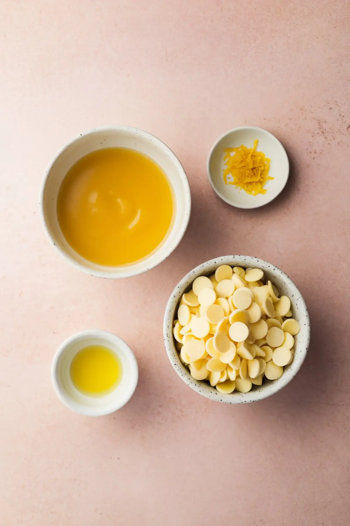 Flat lay image of ingredients for lemon curd truffles, including a bowl of lemon curd, white chocolate chips, lemon zest, and lemon juice in small ceramic dishes on a pastel pink background.