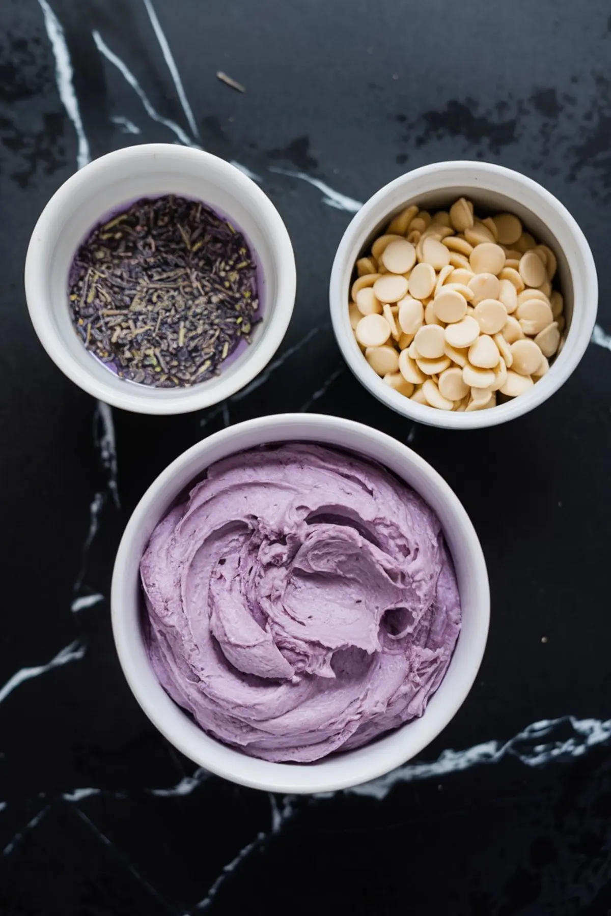 Overhead view of three bowls containing dried lavender soaked in milk, white chocolate chips, and creamy purple lavender-infused butter for bread filling.