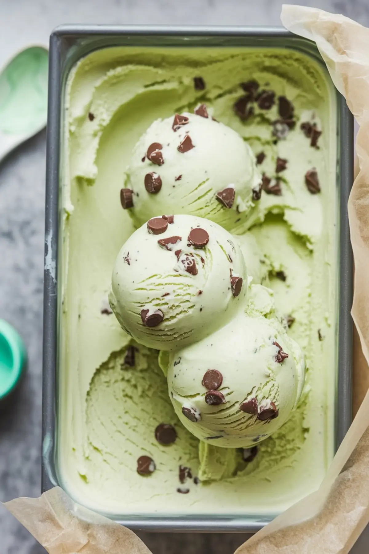 A rectangular metal container holding scoops of mint chocolate chip ice cream, garnished with chocolate chips, with smooth swirls showing the creamy texture.
