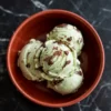 Three scoops of mint chocolate chip ice cream in a terracotta bowl, with visible chocolate shavings and chunks, placed on a black marble surface.