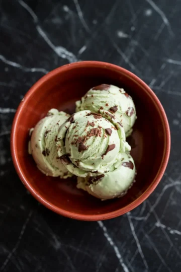 Three scoops of mint chocolate chip ice cream in a terracotta bowl, with visible chocolate shavings and chunks, placed on a black marble surface.