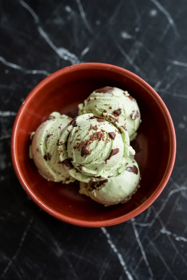 Three scoops of mint chocolate chip ice cream in a terracotta bowl, with visible chocolate shavings and chunks, placed on a black marble surface.