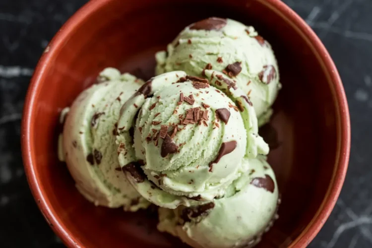 Three scoops of mint chocolate chip ice cream in a terracotta bowl, with visible chocolate shavings and chunks, placed on a black marble surface.