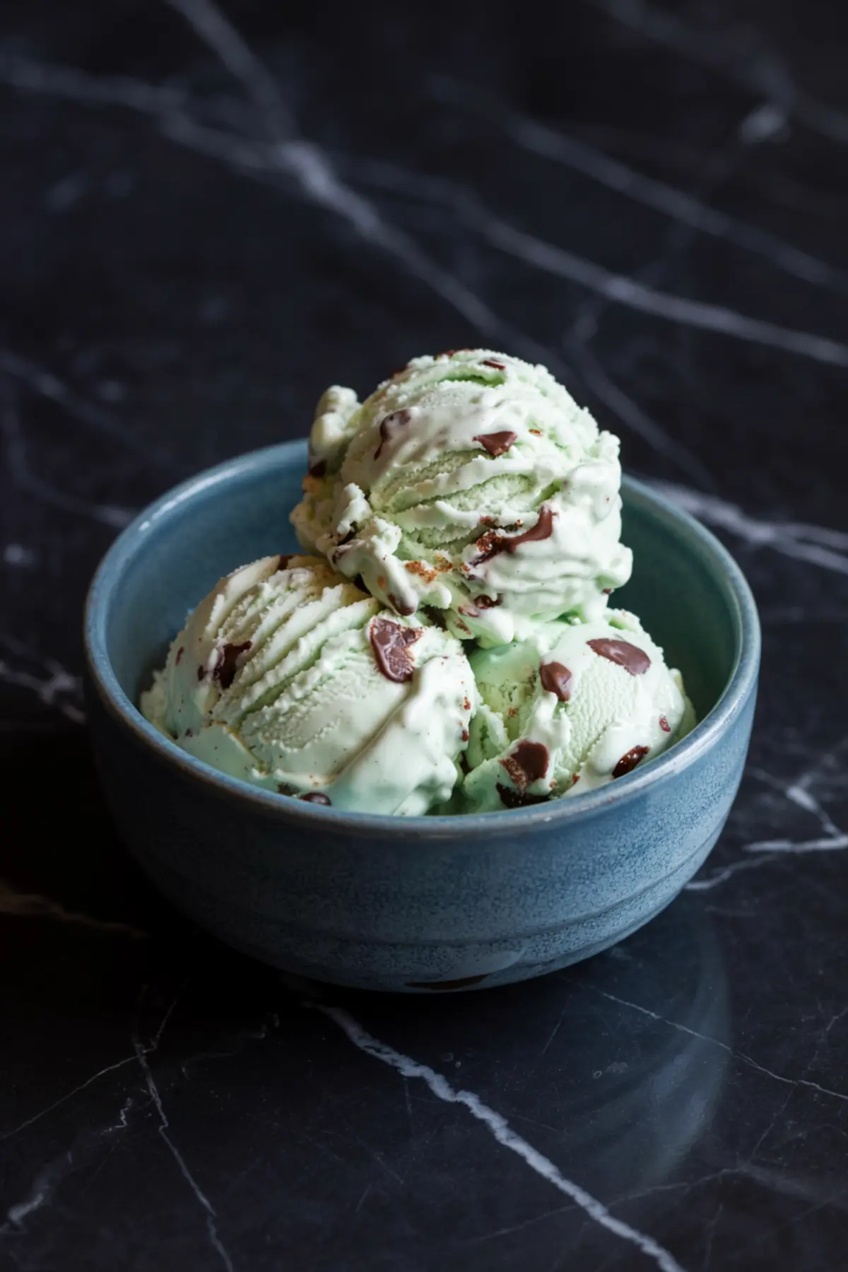 A blue ceramic bowl filled with three scoops of mint chocolate chip ice cream, showing a smooth texture with embedded chocolate bits on a black stone countertop.