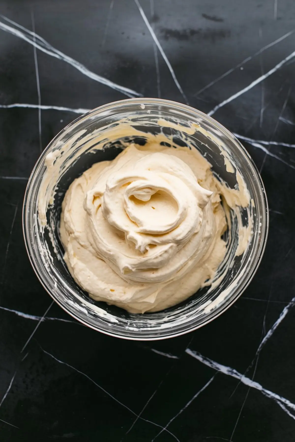 Bowl of whipped cream cheese frosting mixture for fruit pizza, swirled and smooth, displayed on a black marble surface in a glass bowl.