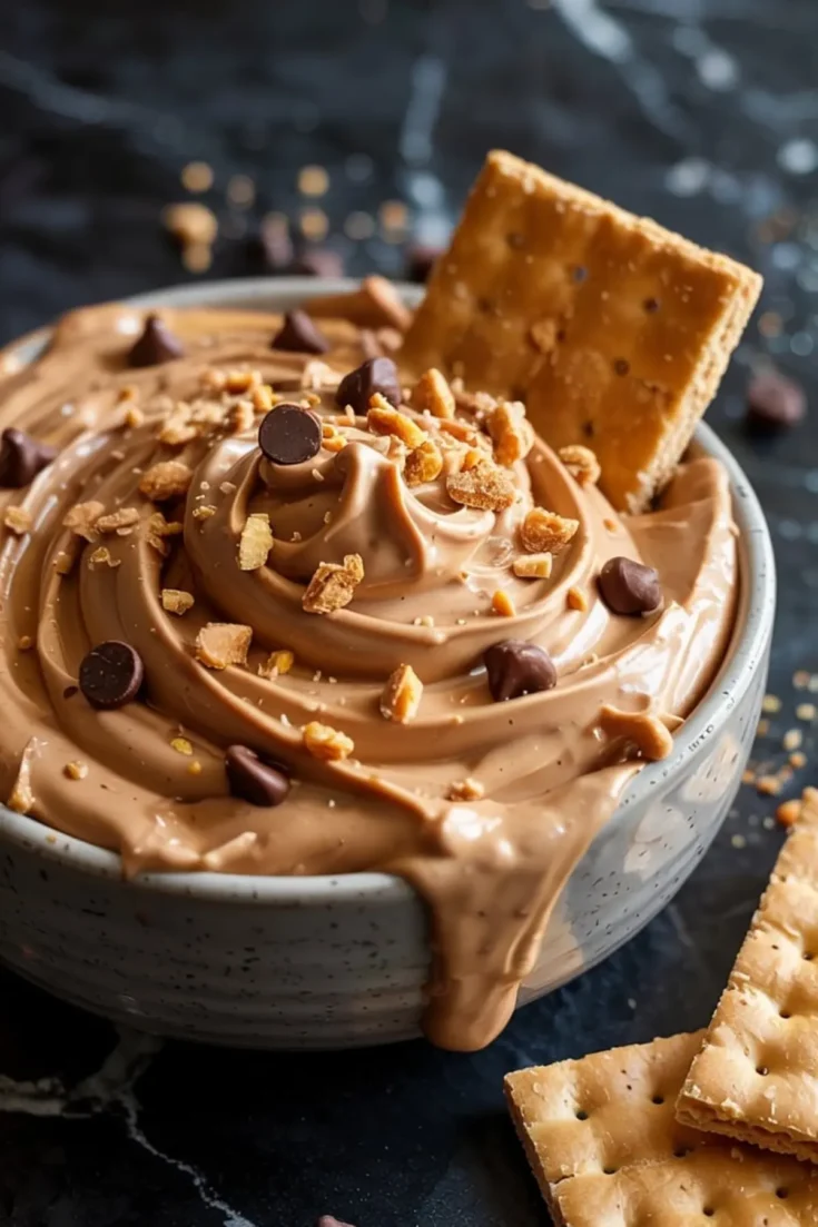Creamy peanut butter cheesecake dip swirled in a ceramic bowl, topped with chocolate chips and crushed graham crackers, with graham crackers served on the side for dipping.