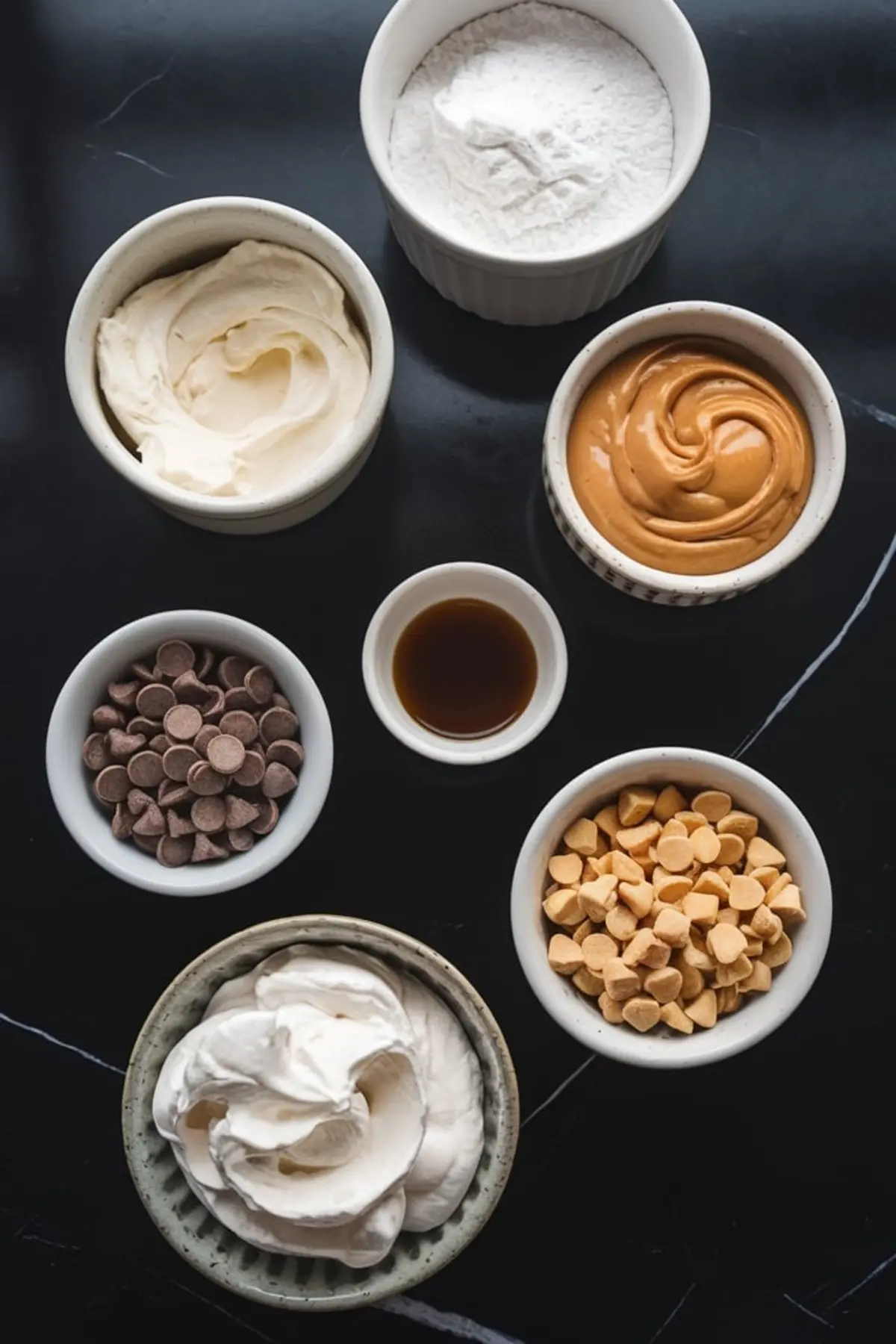 Flat lay of peanut butter cheesecake dip ingredients in small bowls, including cream cheese, powdered sugar, peanut butter, vanilla extract, chocolate chips, peanut butter chips, and whipped topping.
