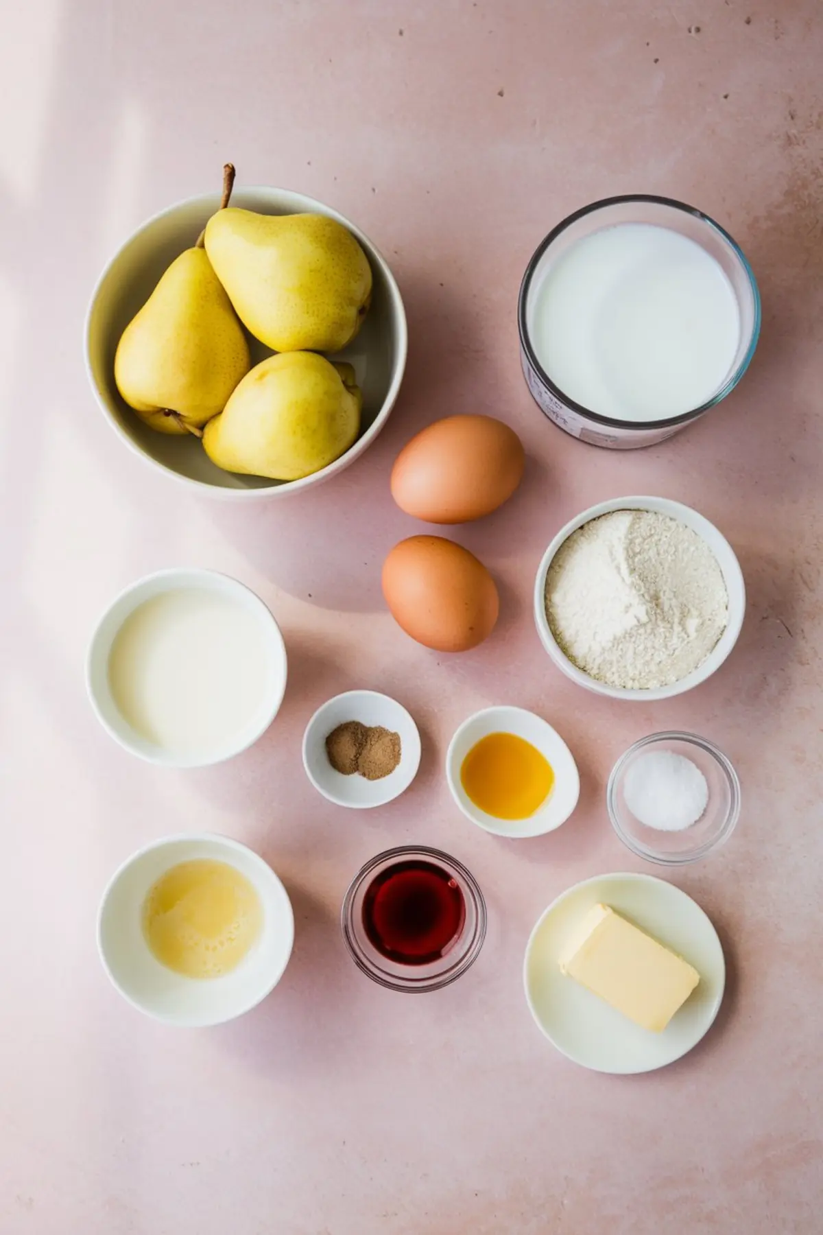Overhead view of ingredients for pear clafoutis arranged on a pink surface, including fresh yellow pears, eggs, milk, flour, butter, sugar, vanilla extract, cream, and warm spices in small bowls.