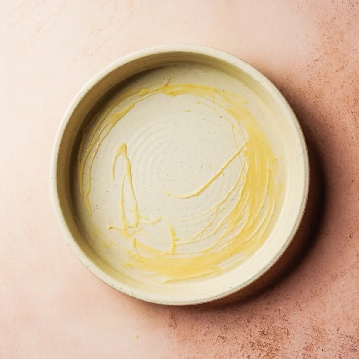 Empty round ceramic baking dish lightly greased with melted butter, set on a pink surface, prepared for pouring in pear clafoutis batter.