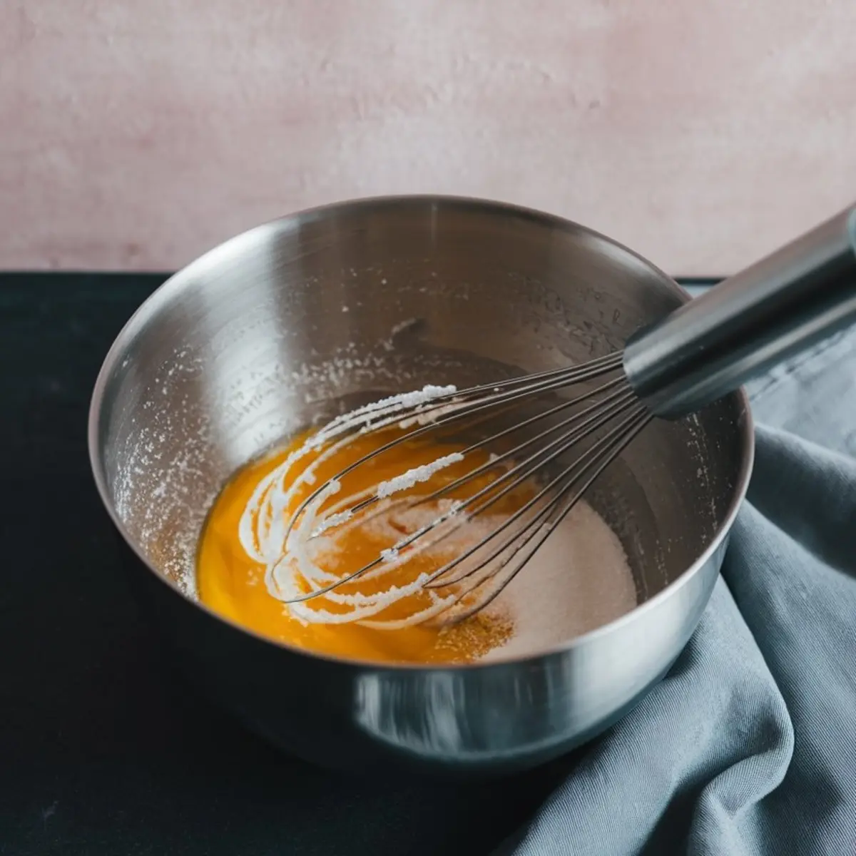Metal mixing bowl with a balloon whisk in the process of blending eggs and sugar into a smooth base for pear clafoutis batter, resting on a soft blue cloth.