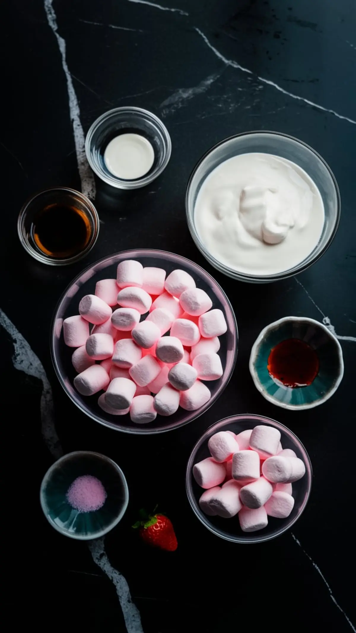 Overhead view of ingredients for pink cloud mousse including pink marshmallows, whipped cream, vanilla extract, strawberry flavoring, cream, and pink powdered gelatin, arranged on a dark marble surface.
