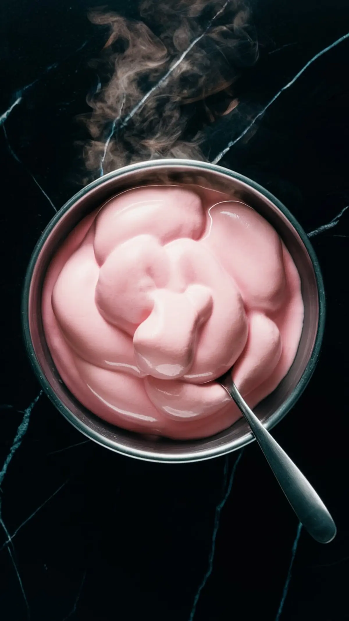 Bowl of freshly melted pink marshmallow mixture for cloud mousse, with visible steam and smooth, creamy consistency, captured against a dark marble background with a silver spoon in the bowl.

