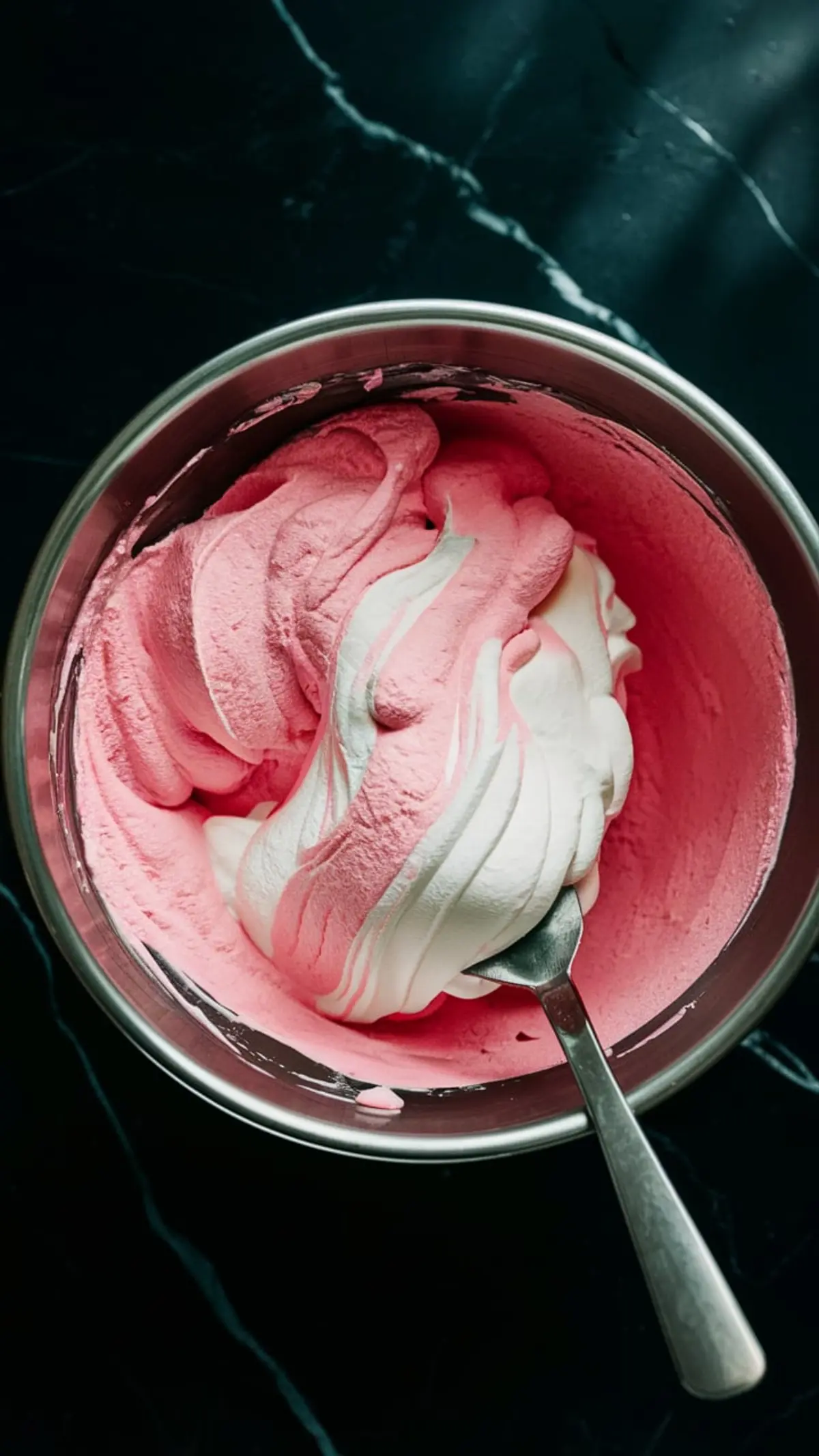 Mixing bowl with swirled pink and white whipped cream in the process of being folded together, showing a light, fluffy texture ideal for creating cloud mousse, photographed on a dark countertop.